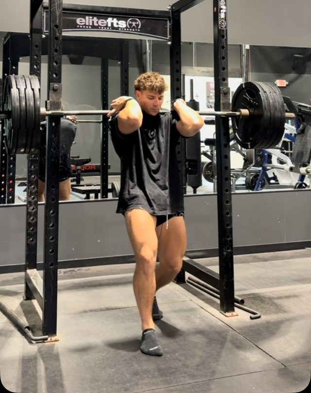 CJ Fazio holding a barbell in the front-rack position, elbows high